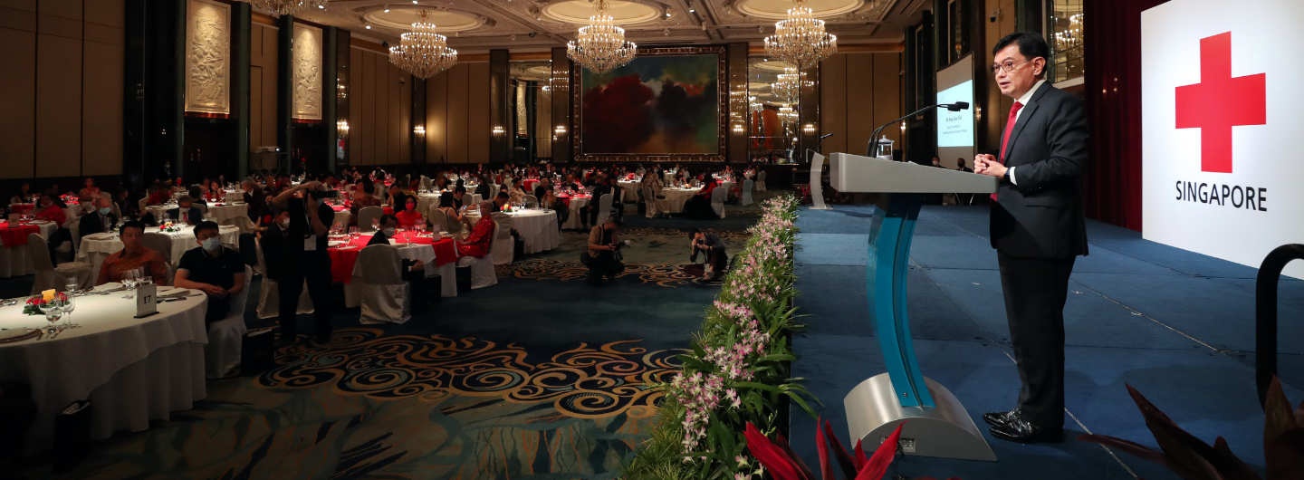 Man at lectern with Red Cross Singapore logo, seated crowd in formal attire.